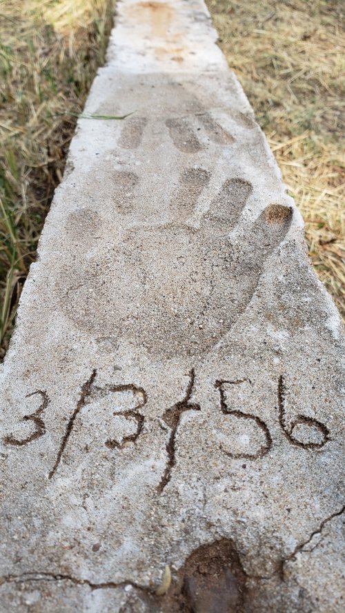 Handprint and date engraved on a concrete slab, surrounded by grass, depicting a natural outdoor setting.
