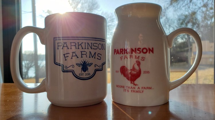 Two ceramic mugs from Parkinson Farms, showcasing blue and red designs with the text "Parkinson Farms" and the establishment year "Est 2016." The blue mug features a bee graphic, while the red jug includes a rooster illustration, all set against a bright, sunlit window backdrop.
