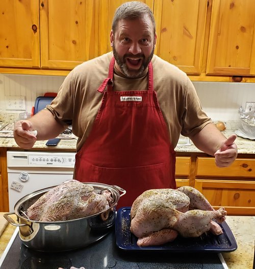 Man in a red apron smiling and giving thumbs up while preparing two seasoned turkeys in a kitchen setting with wooden cabinets and countertops.