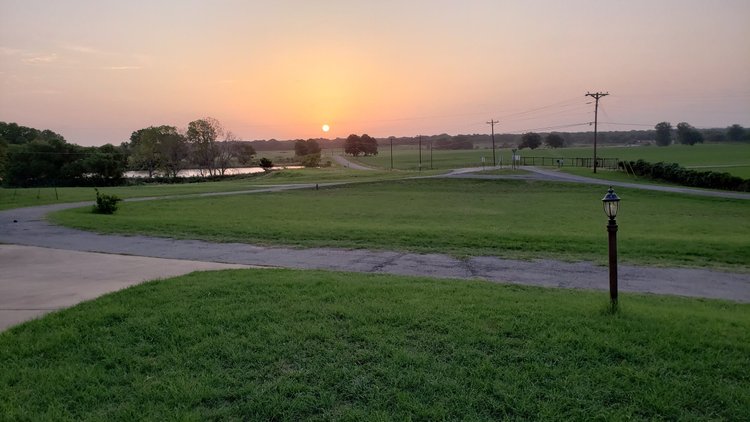Scenic view of a sunset over a rural landscape, featuring lush green grass, a winding road, and a tranquil pond, with utility poles in the background.