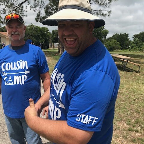Two men wearing blue "Cousin Camp" t-shirts stand outdoors, one smiling and showing his "STAFF" designation, while the other poses with a thumbs up. The setting features green grass and trees, indicating a summer camp environment.