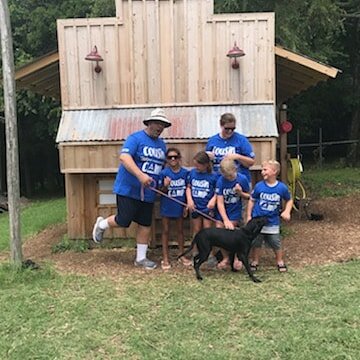 Group of children and adults wearing matching blue "Cousin Camp" shirts, posing in front of a wooden playhouse, with a black dog in the foreground, set in a grassy outdoor area.