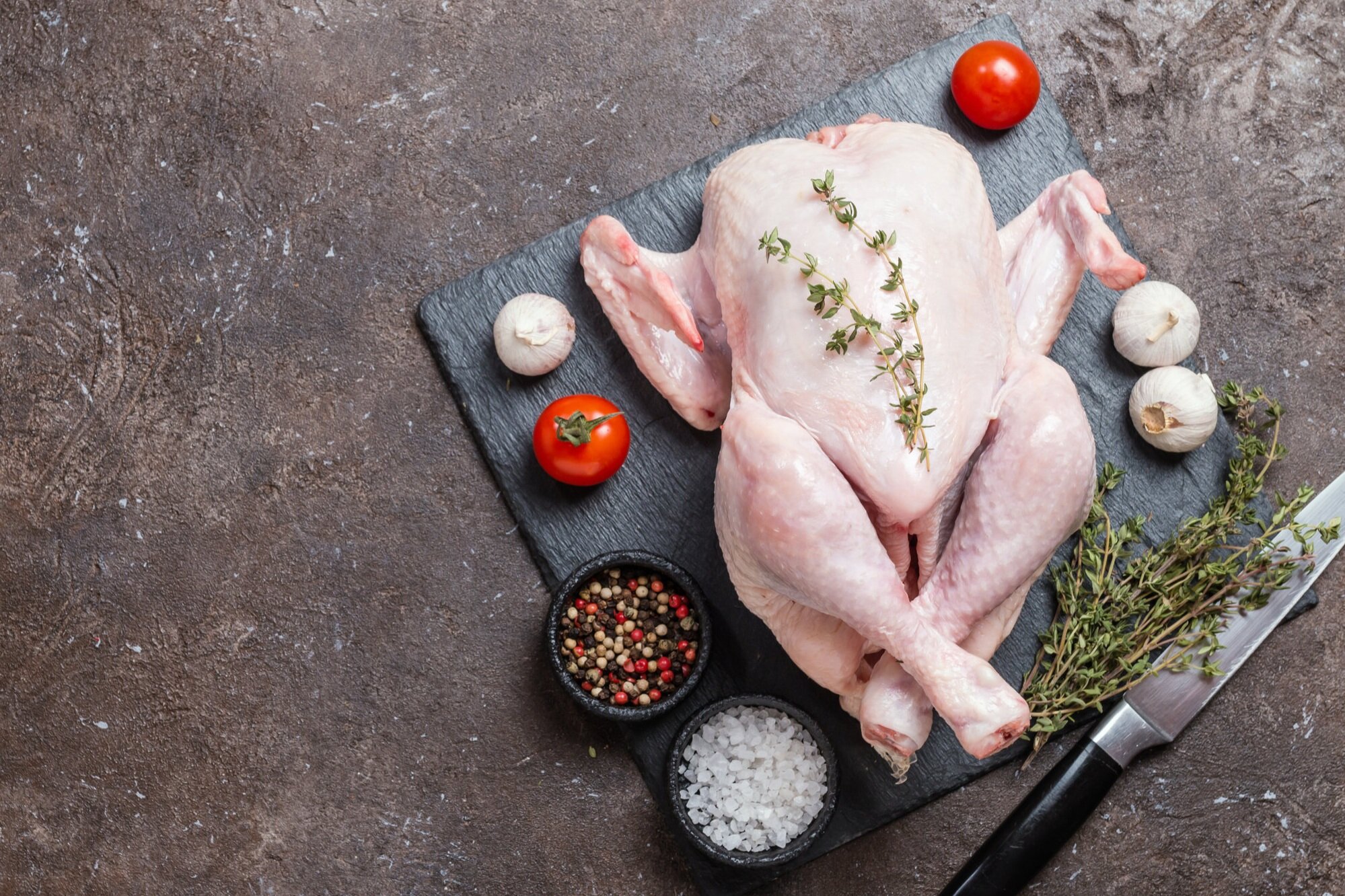 Raw whole chicken with fresh thyme, garlic, and tomatoes on a slate cutting board, accompanied by bowls of mixed peppercorns and sea salt, ready for cooking.