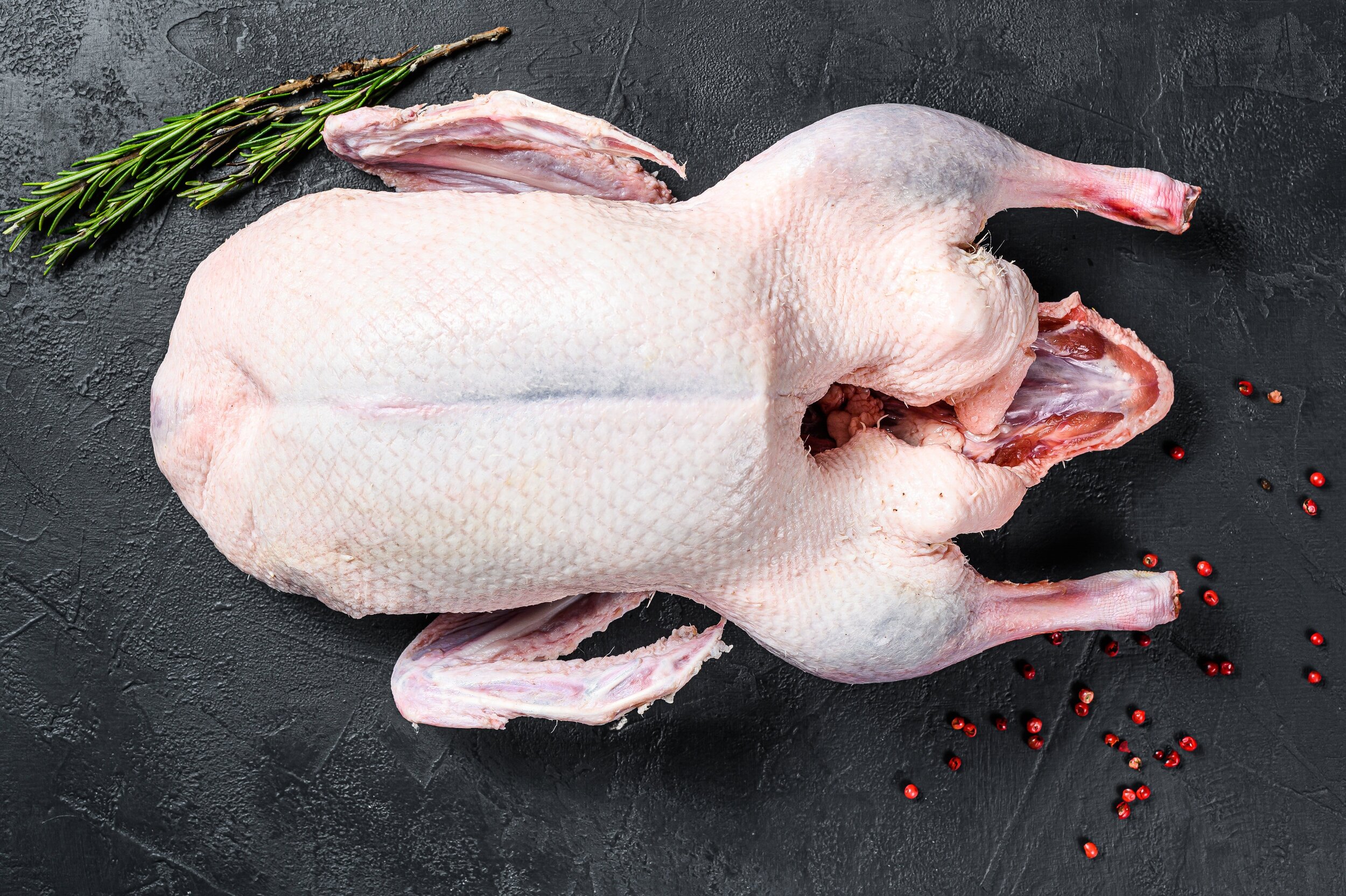 Raw whole duck on a dark textured surface, surrounded by pink peppercorns and a sprig of rosemary, ideal for culinary presentations and recipes.